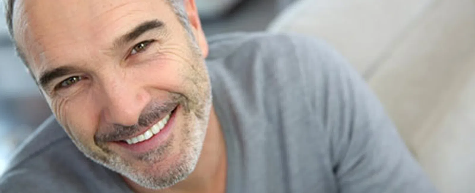 Smiling middle-aged man with gray hair and beard wearing a gray sweater looking at the camera indoors.