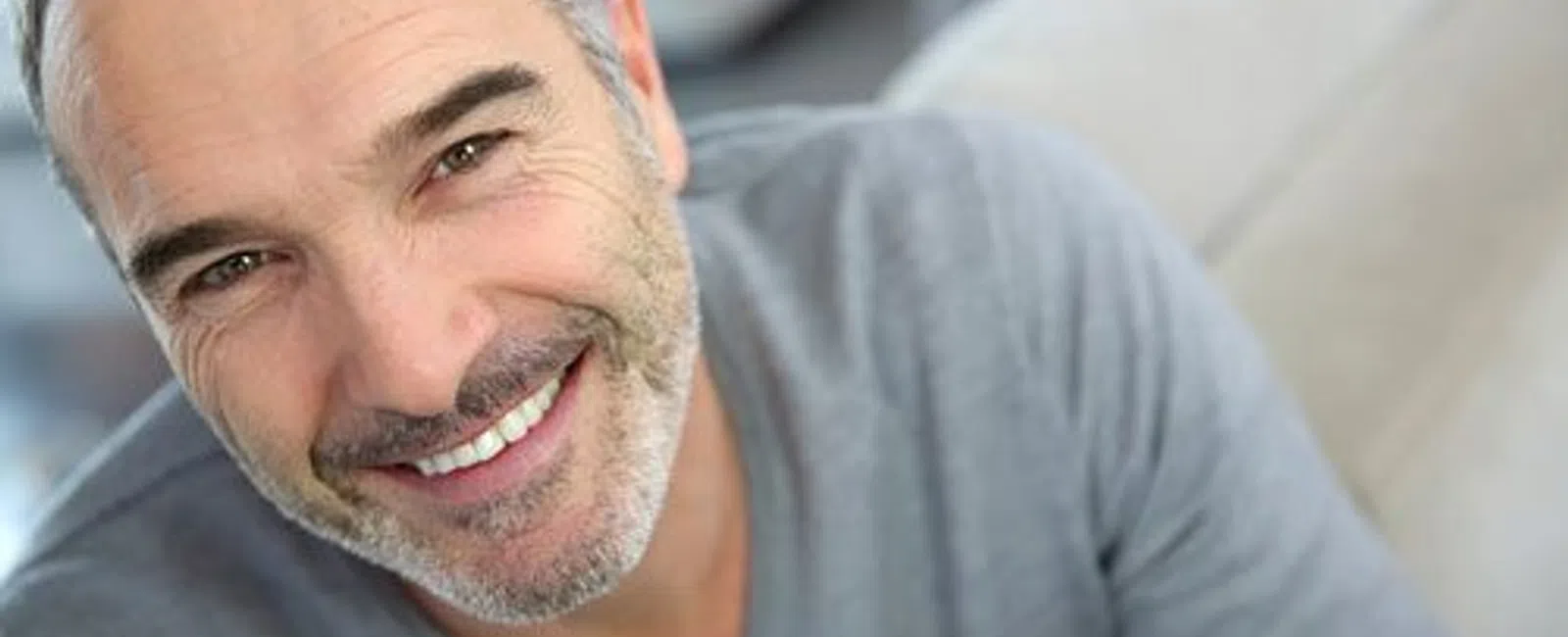 Smiling middle-aged man with gray hair and beard wearing a gray sweater, looking warmly at the camera indoors.