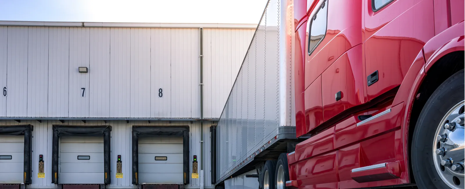 Red semi truck parked at warehouse loading dock with numbered doors under clear sky