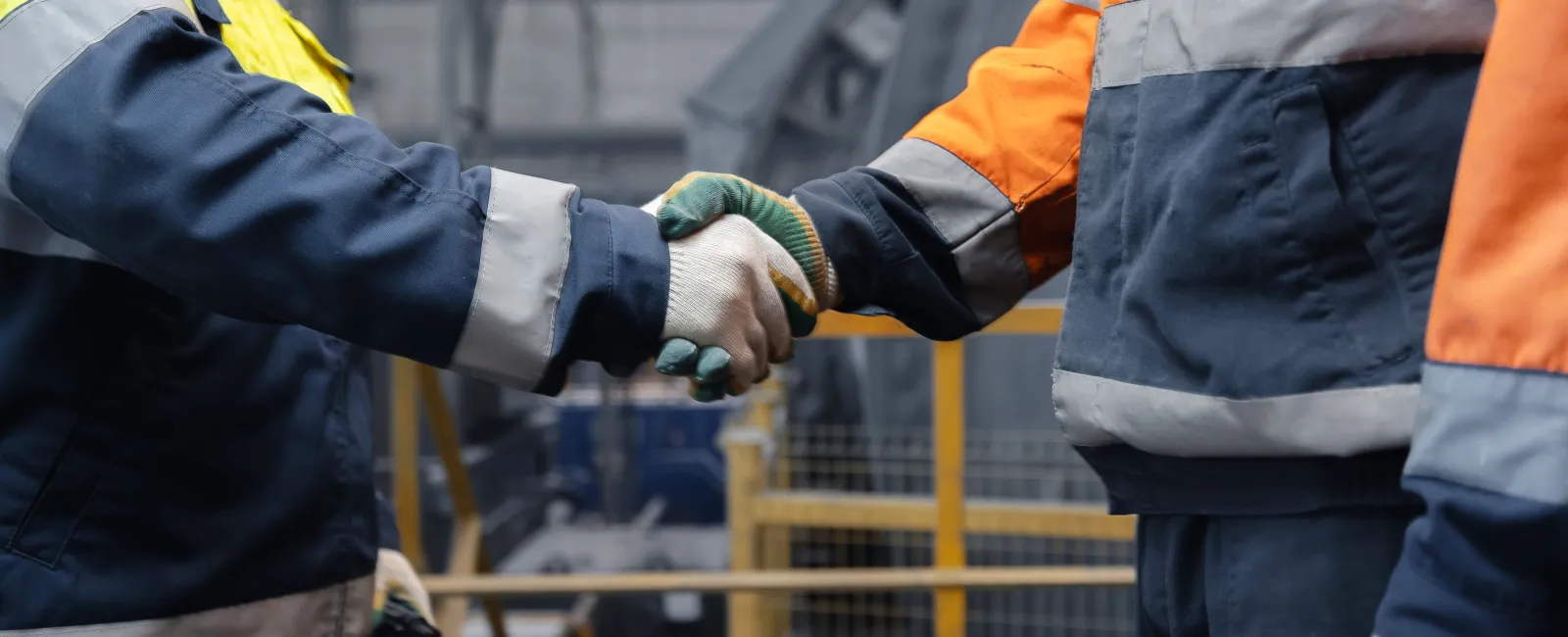 Two industrial workers wearing safety jackets and gloves shaking hands inside a factory setting.