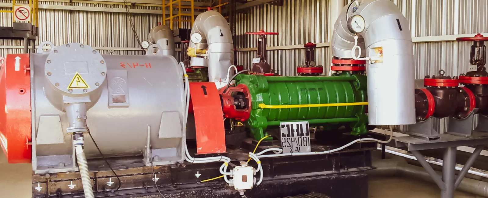 Industrial pump machinery with green and metallic components inside a factory building with yellow crane overhead.