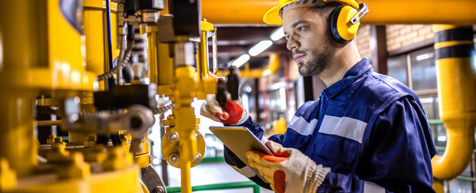 Industrial worker in safety gear inspecting machinery and taking notes on a tablet in a factory setting