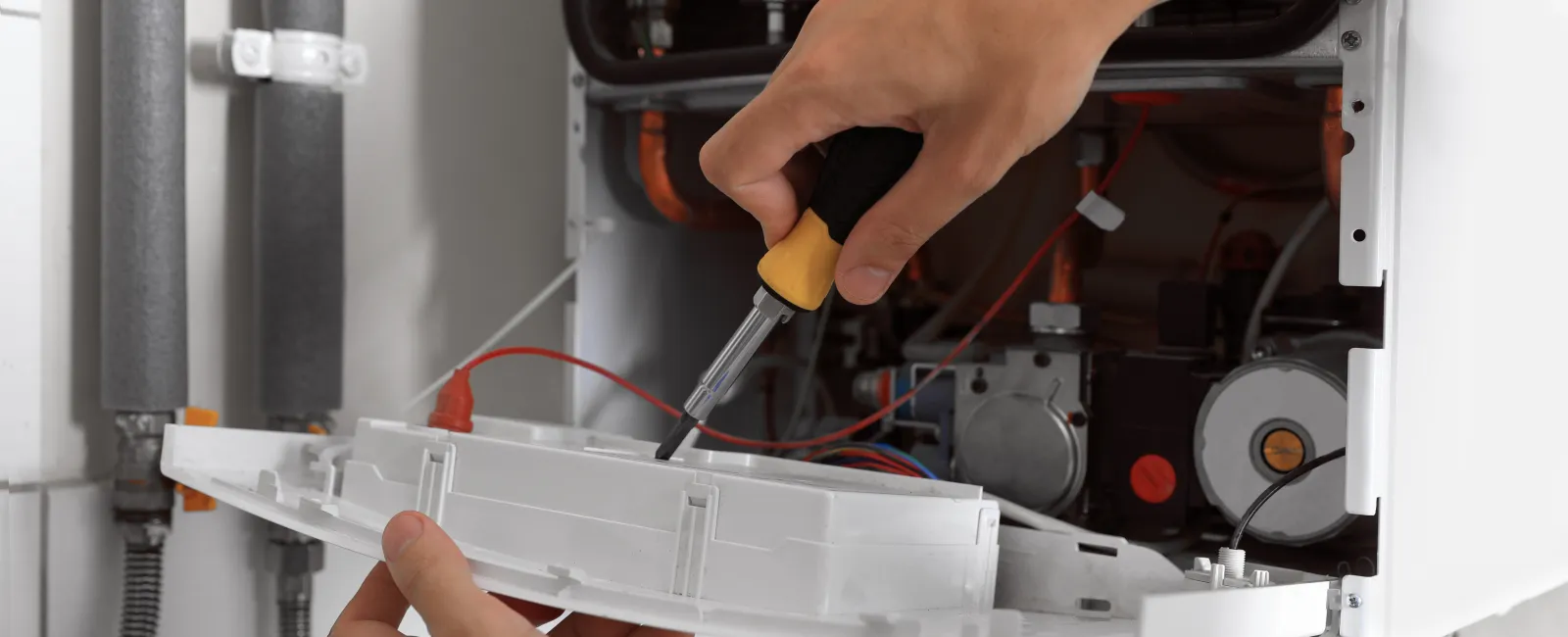 Technician repairing a boiler using a screwdriver with the boiler panel removed showing internal components.