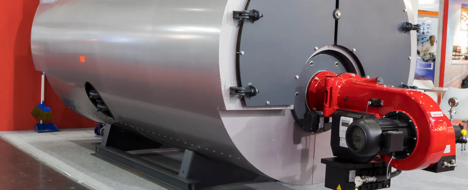 Large industrial boiler with red burner and metal body displayed indoors at trade show.