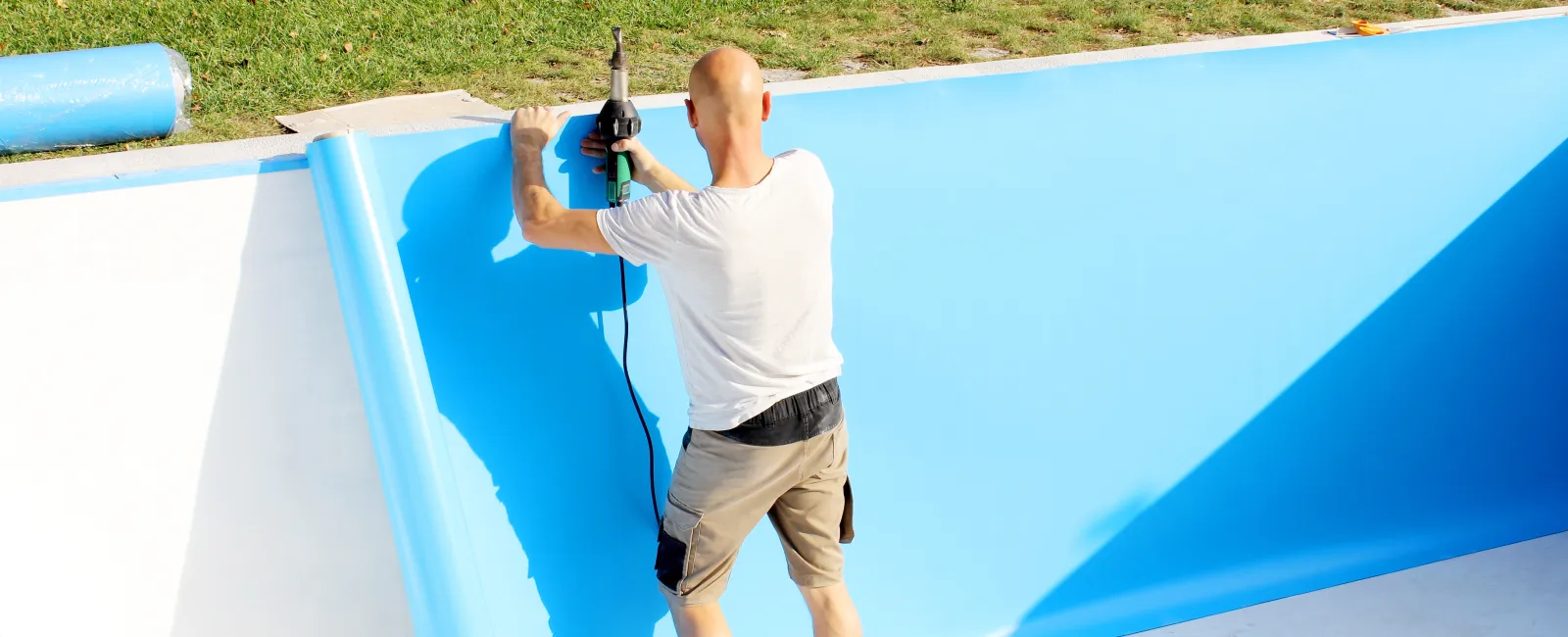 Man installing blue pool liner using a power tool inside an empty swimming pool on a sunny day