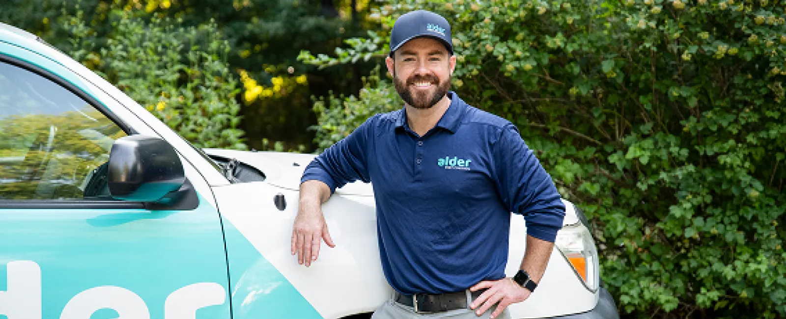 Alder Control technician poses beside a branded service vehicle in a green outdoor setting.