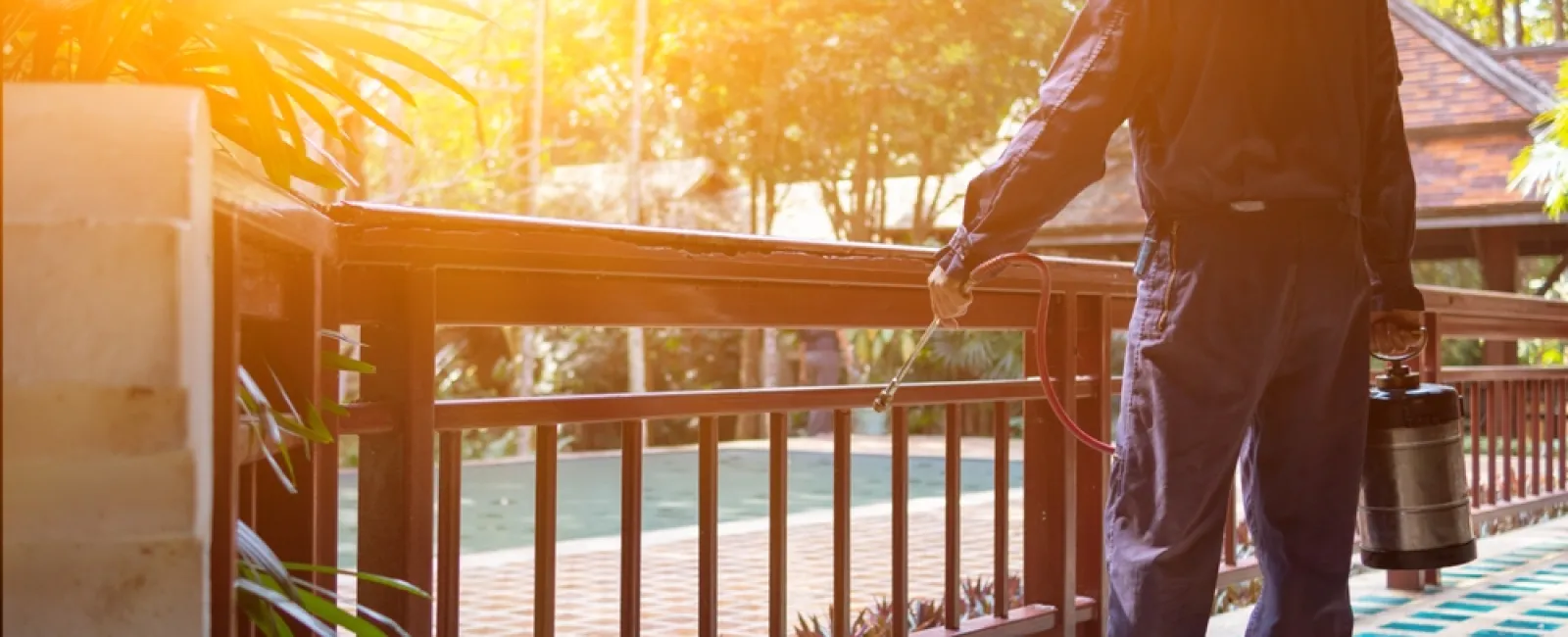 Man in protective clothing spraying insecticide on outdoor wooden railing during daytime.