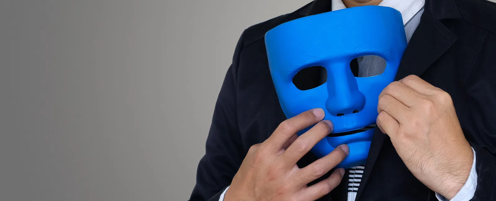 Person in suit holding a blue mask near face against a gray background.