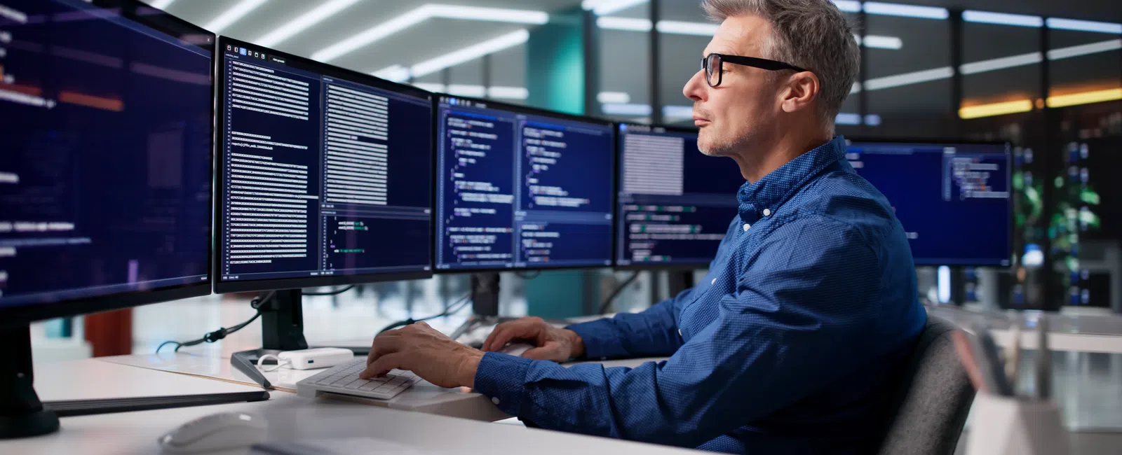 Man working on multiple computer monitors displaying code in a modern office environment.