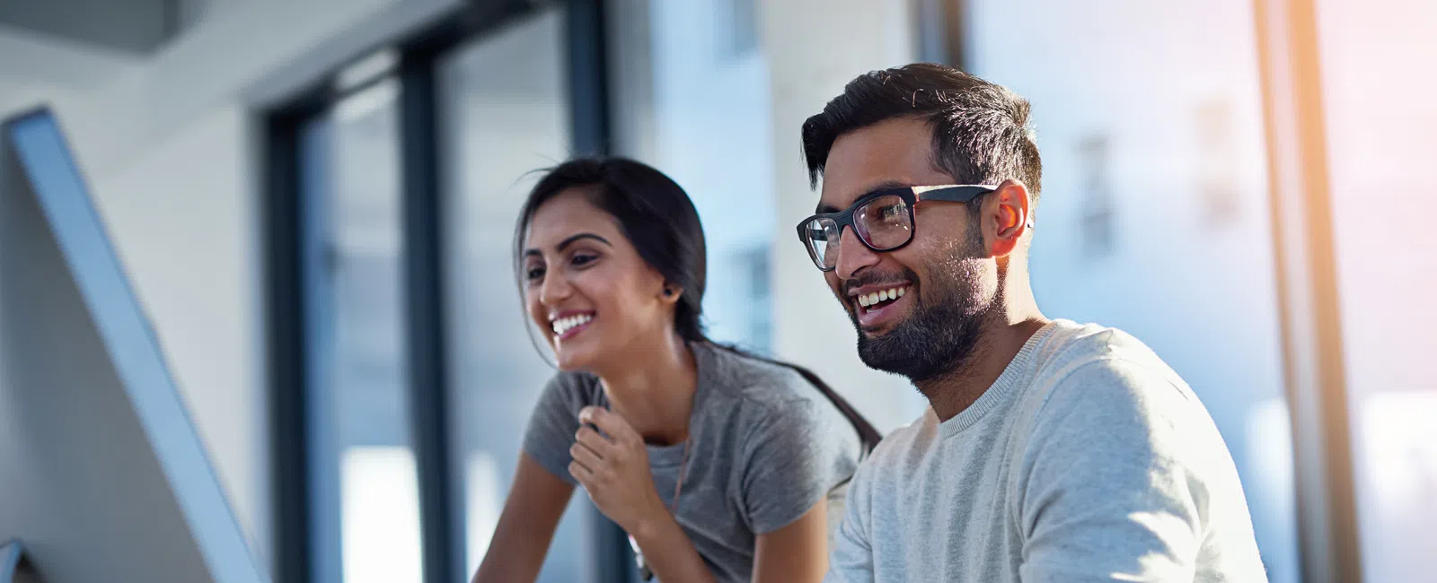 Two coworkers smiling and collaborating while working on a desktop computer in a bright modern office.
