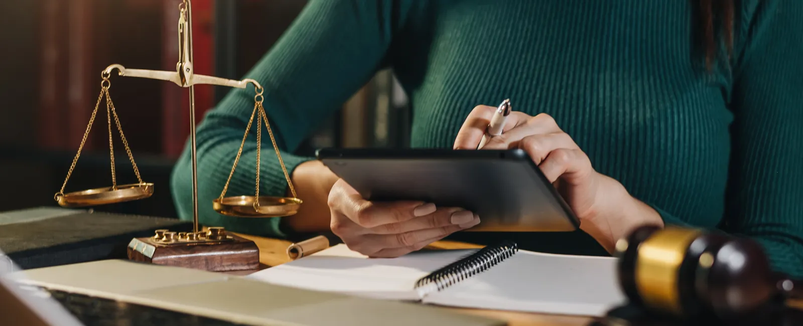 Woman working with tablet, law scales, gavel, and laptop on wooden desk in legal office setting.