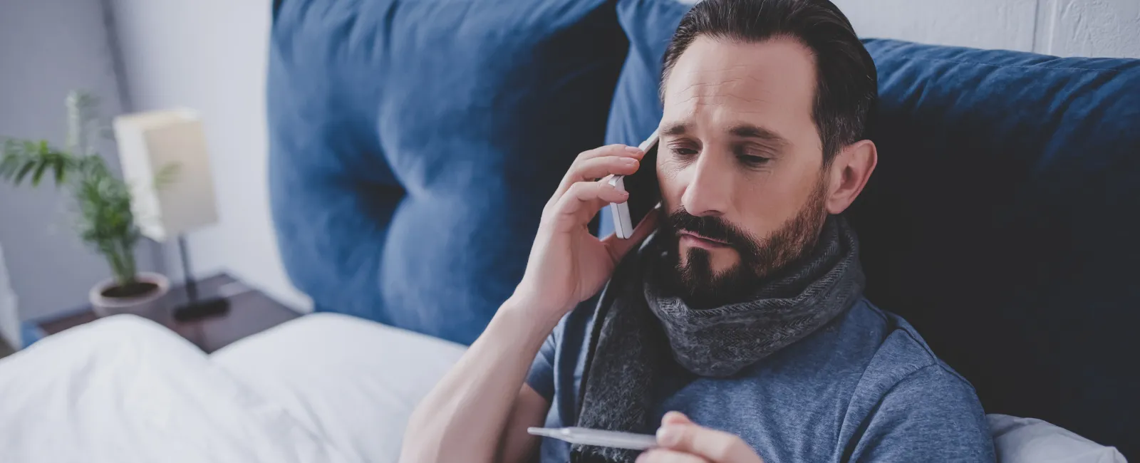 Man in bed wearing scarf checks thermometer while talking on phone, possibly sick and seeking advice.