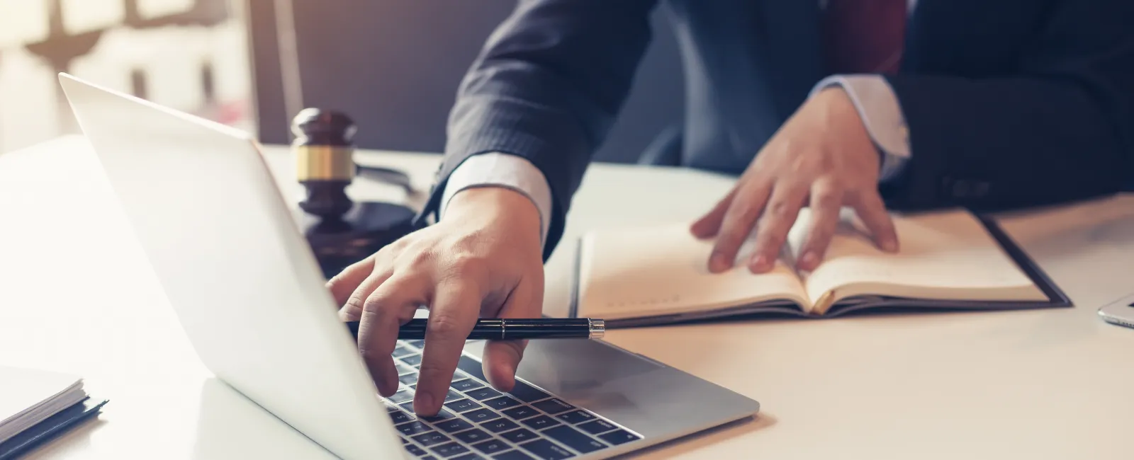Businessman in suit using laptop and taking notes with a pen in office setting with gavel nearby.