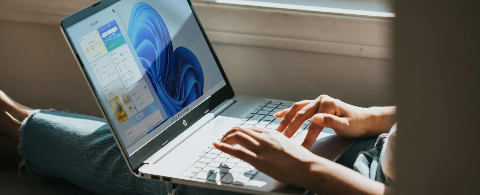 Person typing on a laptop with Windows 11 home screen, sitting cross-legged by a window in natural light.
