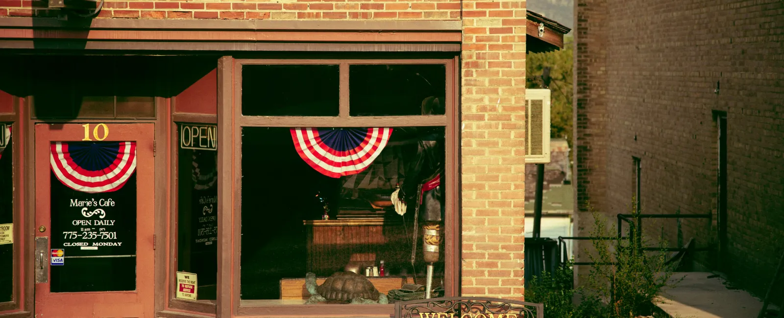 Exterior of a brick rural small business cafe with patriotic red, white, and blue bunting, a welcome bench, and an open sign.