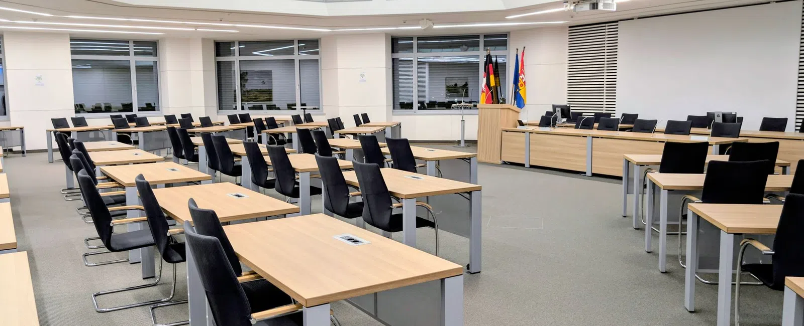 Empty modern conference room with multiple desks, black chairs, flags, and a large skylight ceiling.