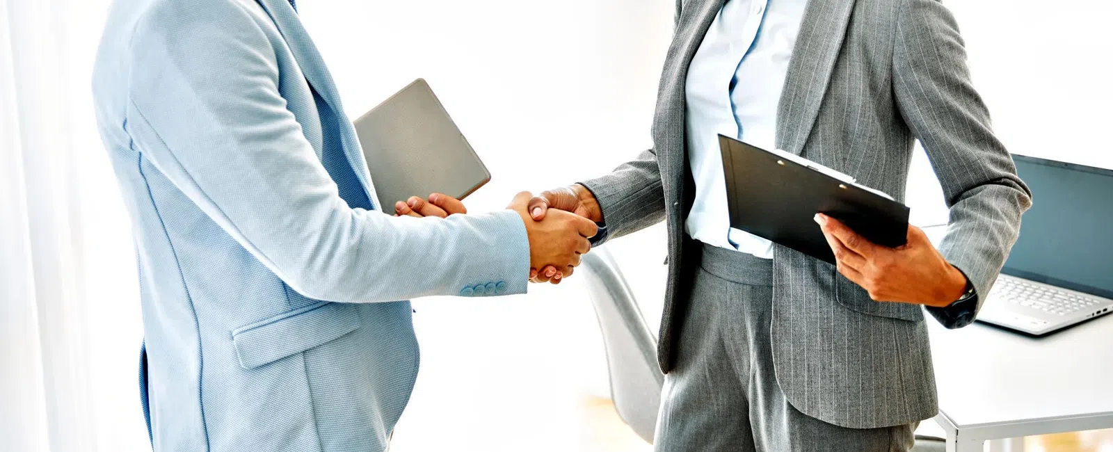 Two business professionals shaking hands in bright office setting, smiling and holding documents during meeting.