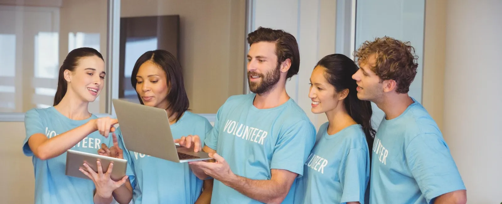 Group of volunteers in blue shirts collaborating using laptop and tablet around a table with colorful notes.