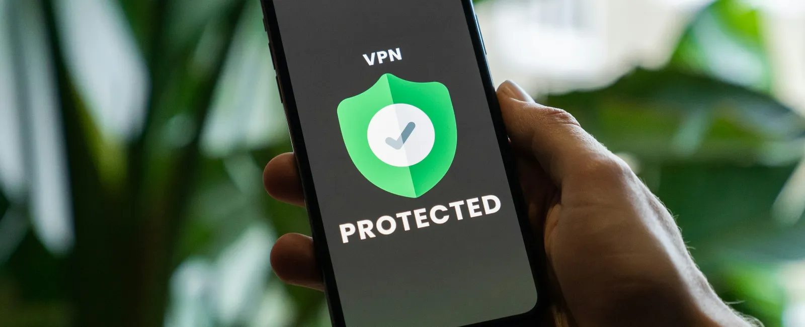 Hand holding smartphone displaying VPN protection shield with checkmark on screen against blurred green background