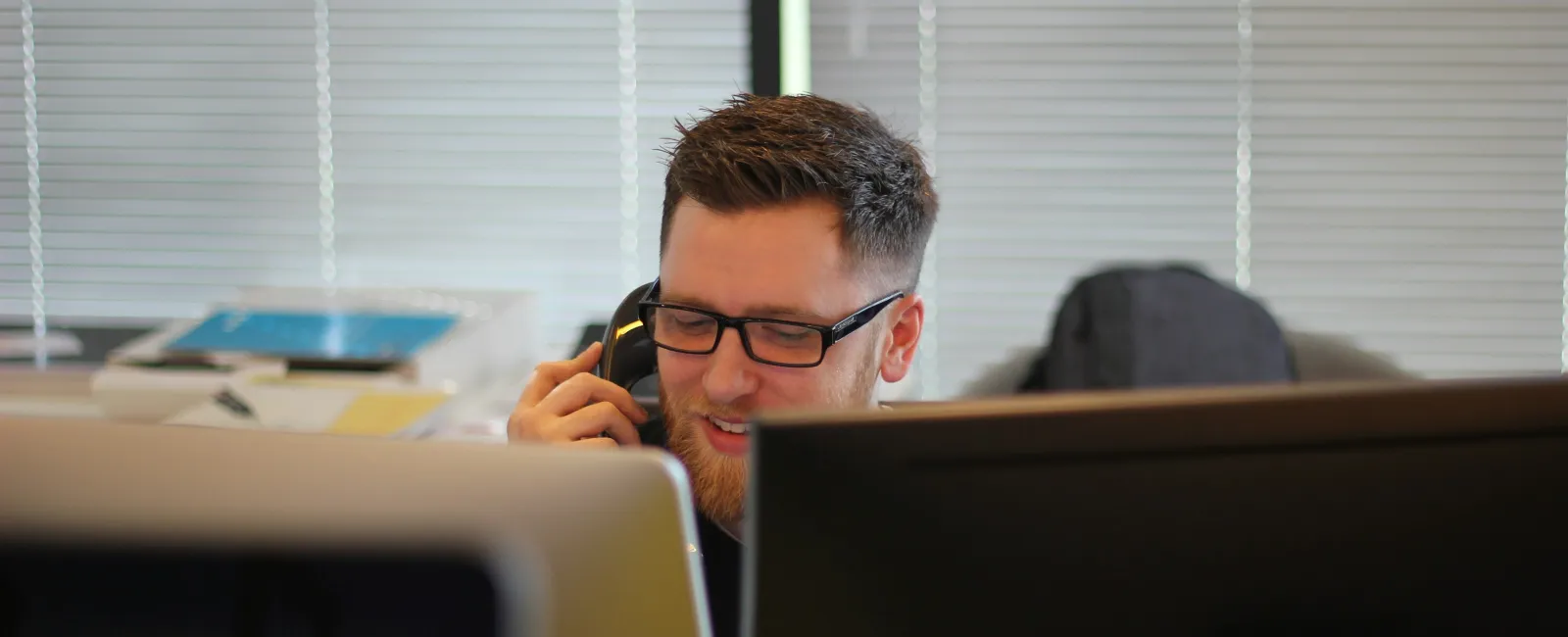 Man wearing glasses talking on phone behind computer monitors in modern office with blinds in background