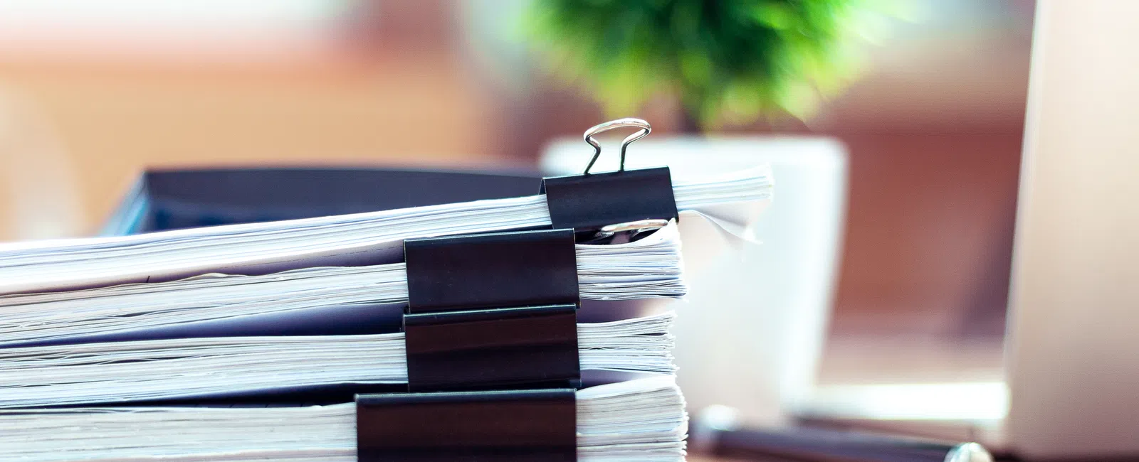 Stack of organized documents with black binder clips on a wooden desk, blurred plant and laptop in background