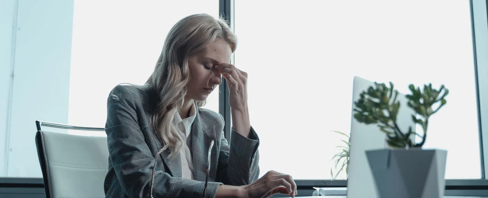 Stressed businesswoman sitting at desk in front of laptop, rubbing forehead in modern office with large windows.
