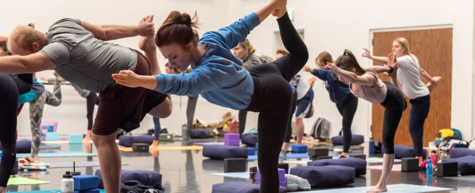 Group yoga class practicing balancing pose in a spacious studio with mats, blocks, and cushions