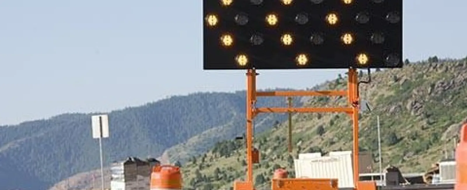 Electronic arrow sign directing traffic right on road near orange construction barrels with mountainous background