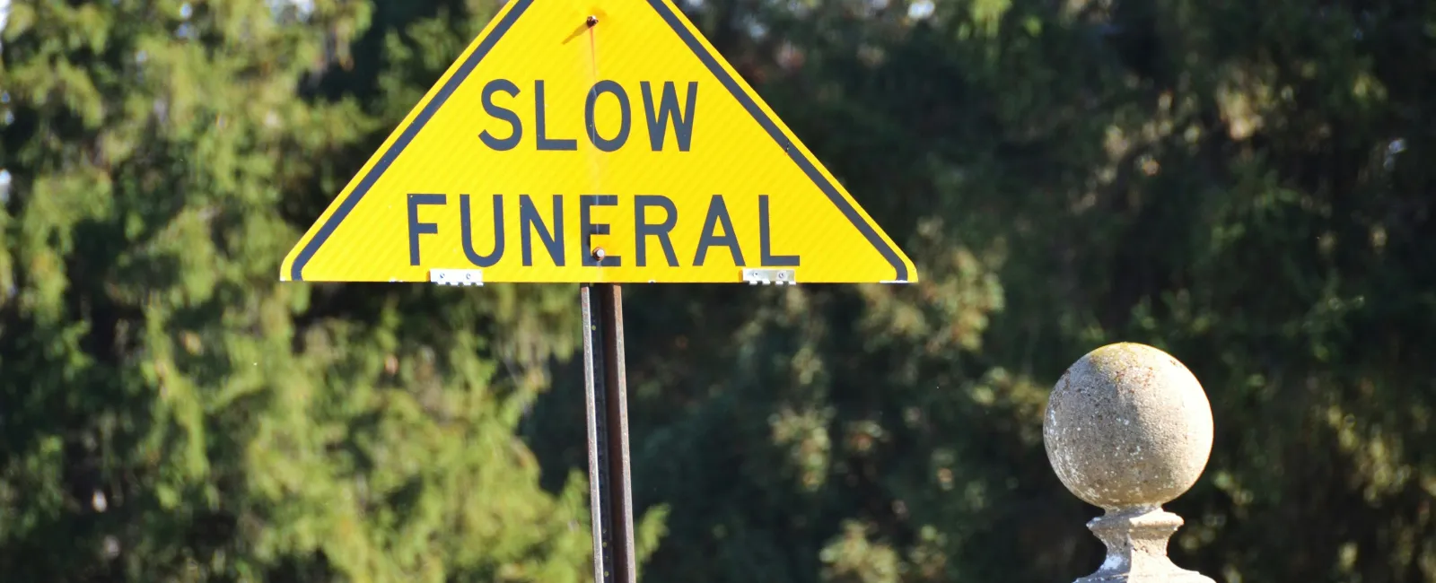 Yellow triangular warning sign reading Slow Funeral near cemetery stone and trees in background