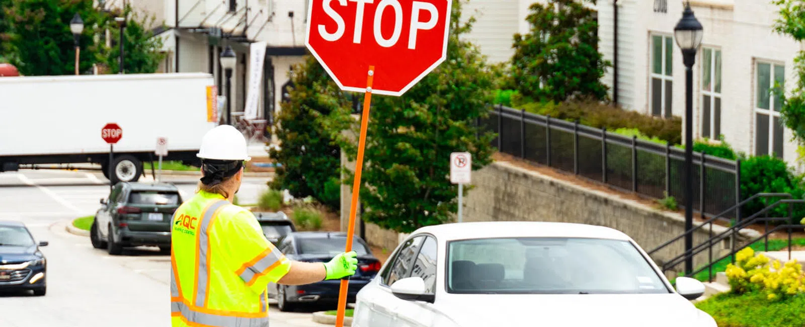 Construction worker in safety gear holding a stop sign directing white car on urban street with buildings.