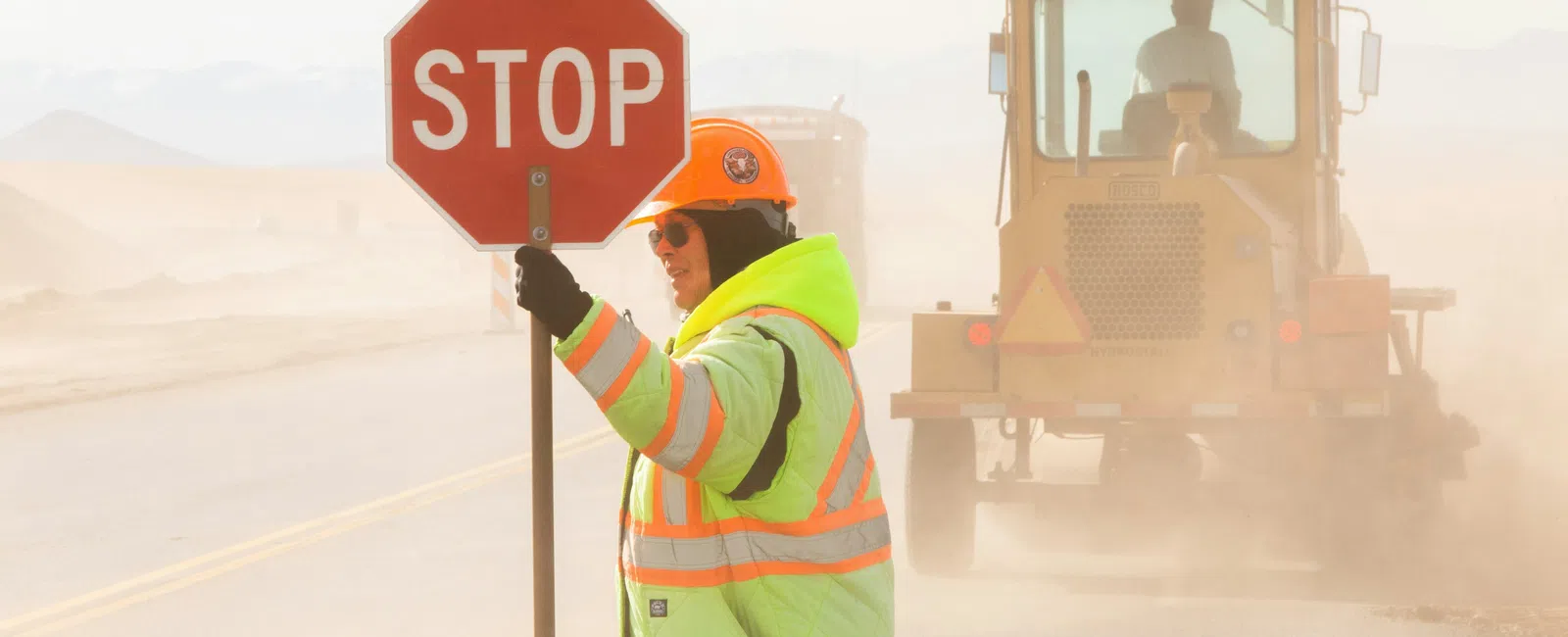 Construction worker in reflective jacket holding stop sign on dusty road with heavy machinery behind.