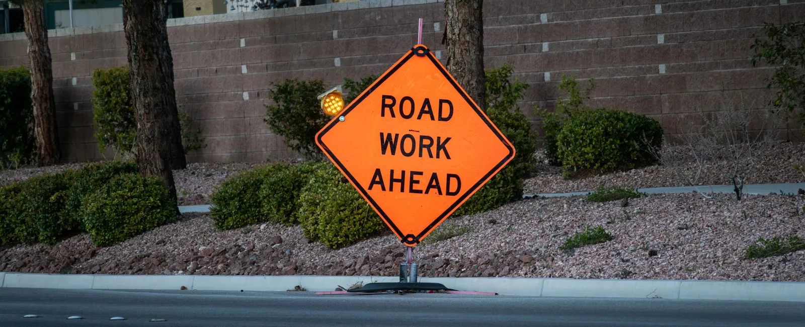 Orange diamond-shaped road sign reading 'Road Work Ahead' placed near street with trees and bushes behind.