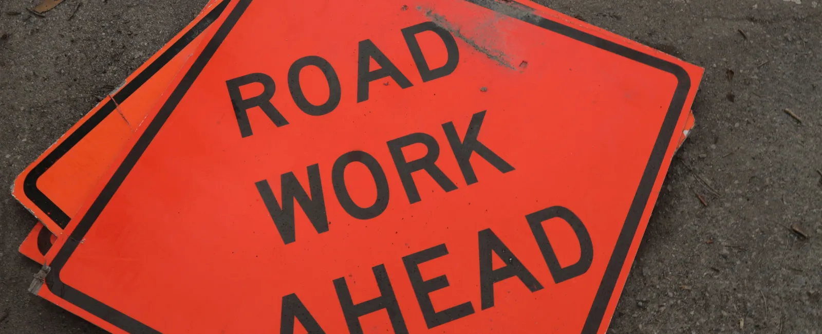 Bright orange road work ahead signs stacked on asphalt surface with black text.
