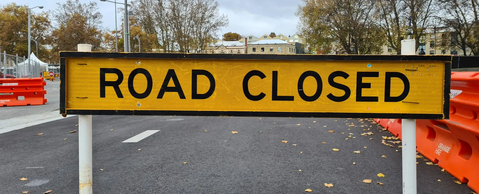 Yellow road closed sign blocking street with orange barriers and trees in the background on a cloudy day