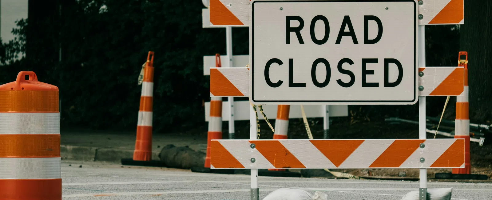 Road closed barrier with orange and white striped traffic barrels blocks street, with overlaid text reading ROAD CLOSED.