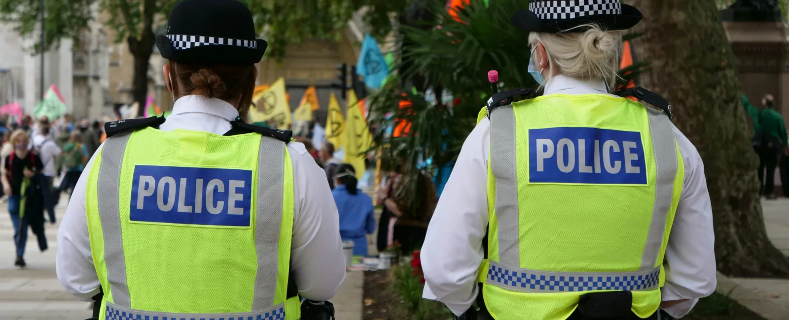 Rear view of two police officers in high-visibility jackets monitoring a crowd at an outdoor event with flags.