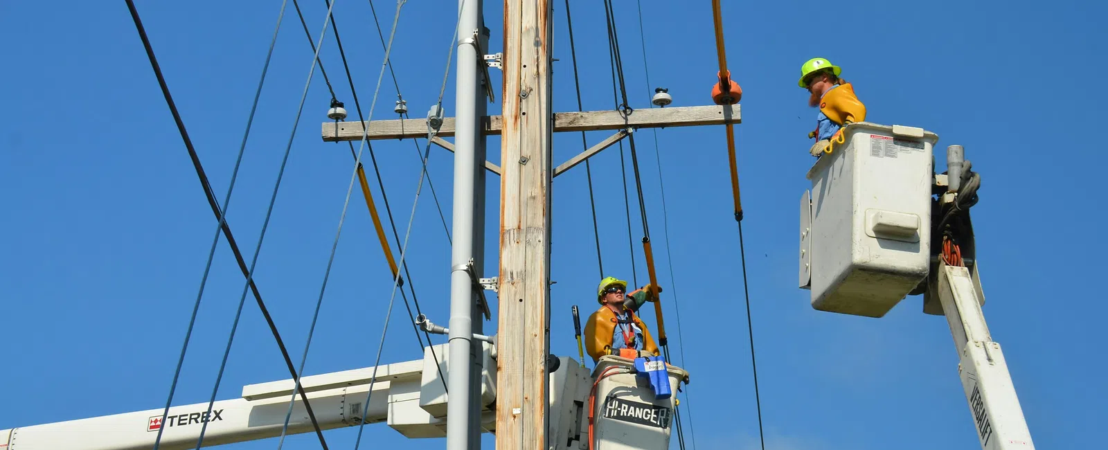 Two linemen in bucket trucks working on a wooden utility pole against a clear blue sky.