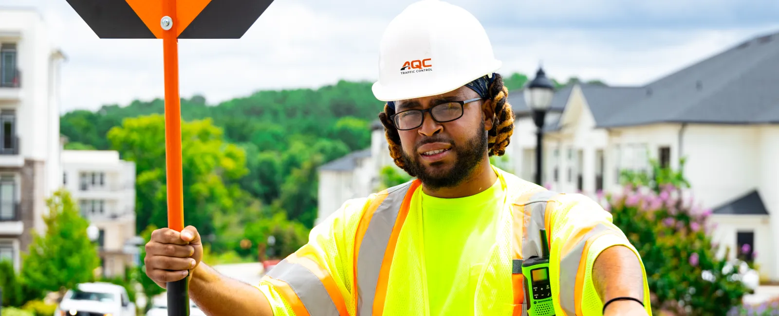 Construction worker wearing safety gear holding slow sign on suburban street with houses and greenery in background