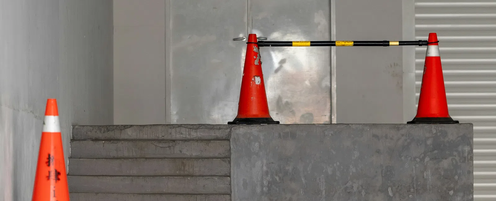 Concrete stairs and platform blocked by orange traffic cones and a horizontal barrier in front of gray metal doors in a parking area