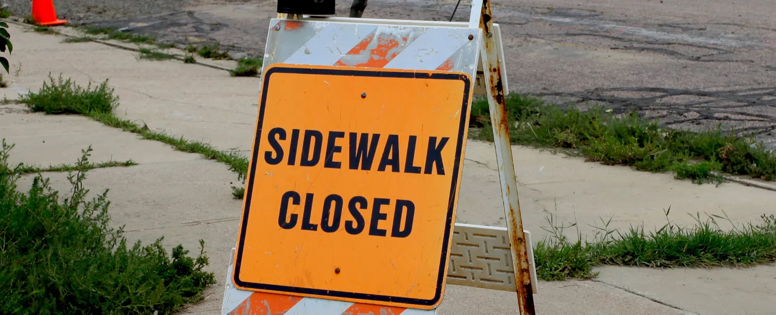 Orange sidewalk closed sign with flashing light blocking cracked sidewalk near street and traffic cones.