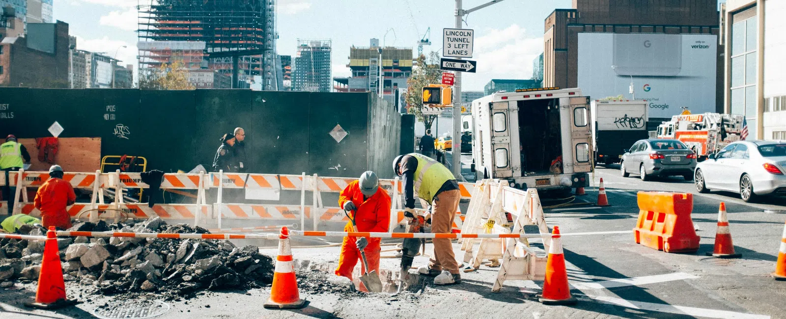 Construction workers repairing urban road with safety cones, barriers, and city buildings in the background.