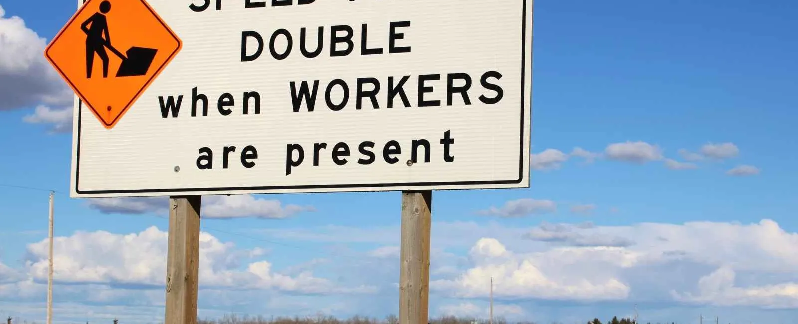 Road sign warning that speed fines double when workers are present, with blue sky and highway in background.