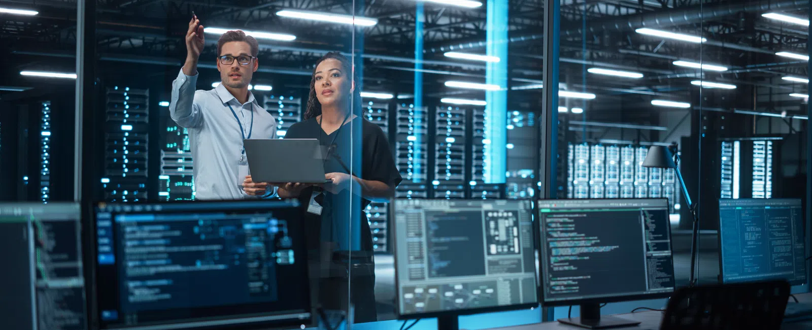 Two IT professionals working on laptops and servers in a modern data center with multiple monitors.