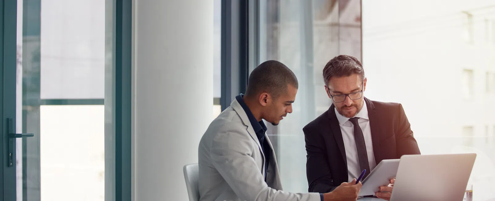 Two businessmen discussing documents at a bright office with laptop and glass of water on the table
