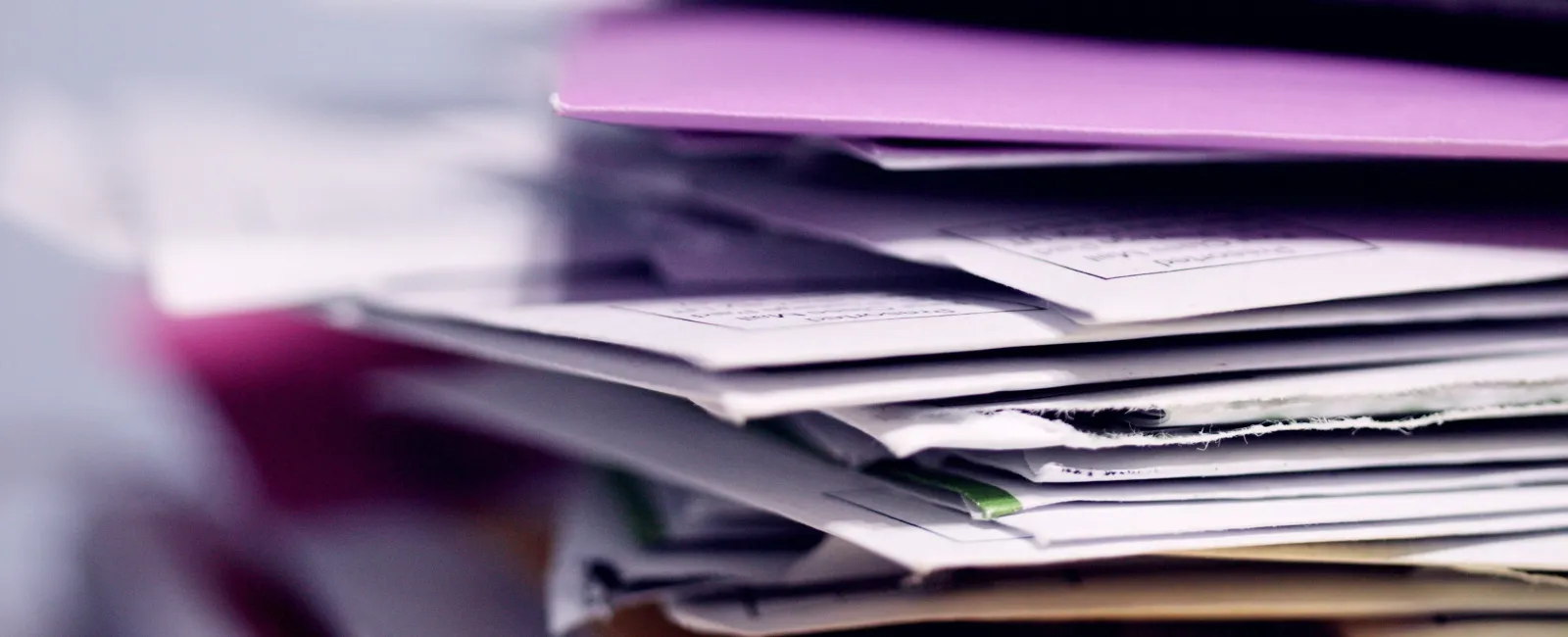 Close-up of a tall stack of colorful envelopes and papers piled messily on a desk surface.
