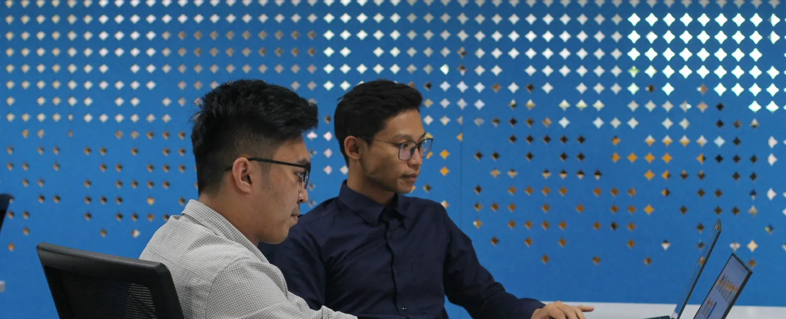 Two men working on laptops at a white desk with a blue perforated partition wall behind them