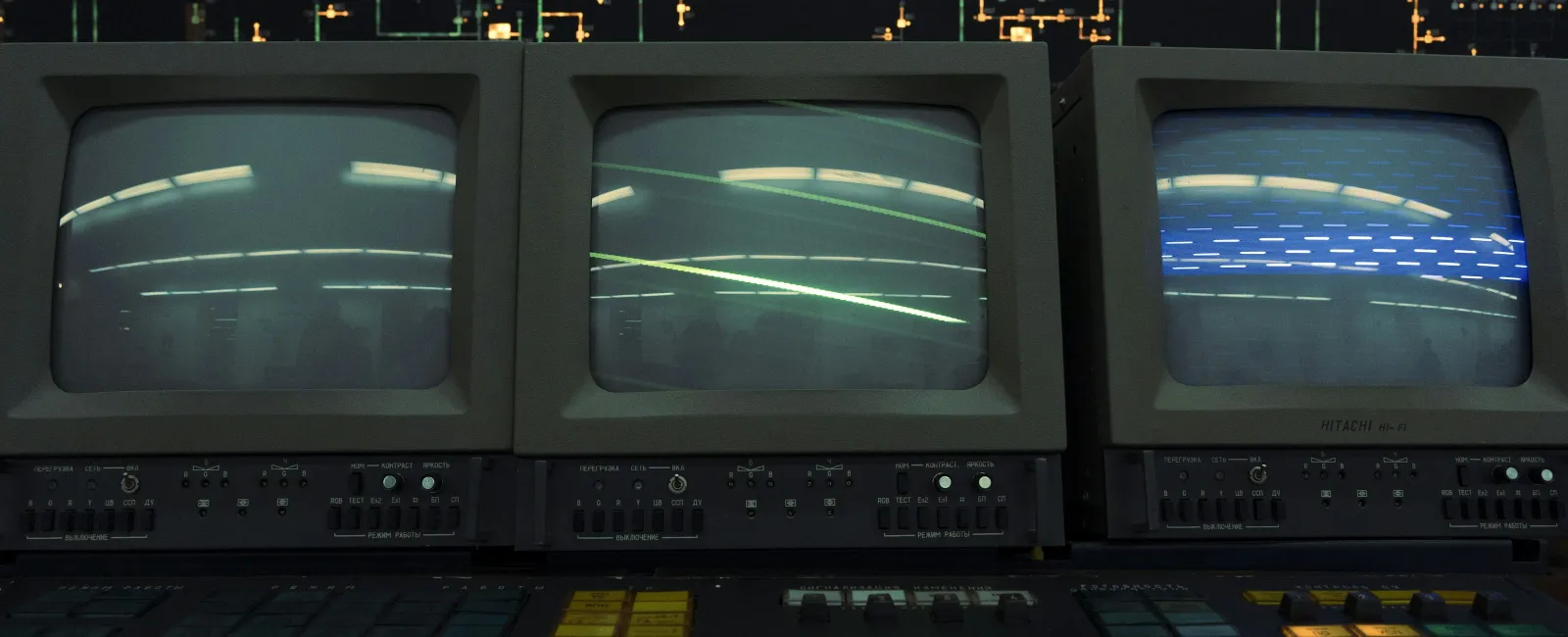 Three vintage CRT monitors displaying static and reflections above a control panel with numerous buttons and switches.