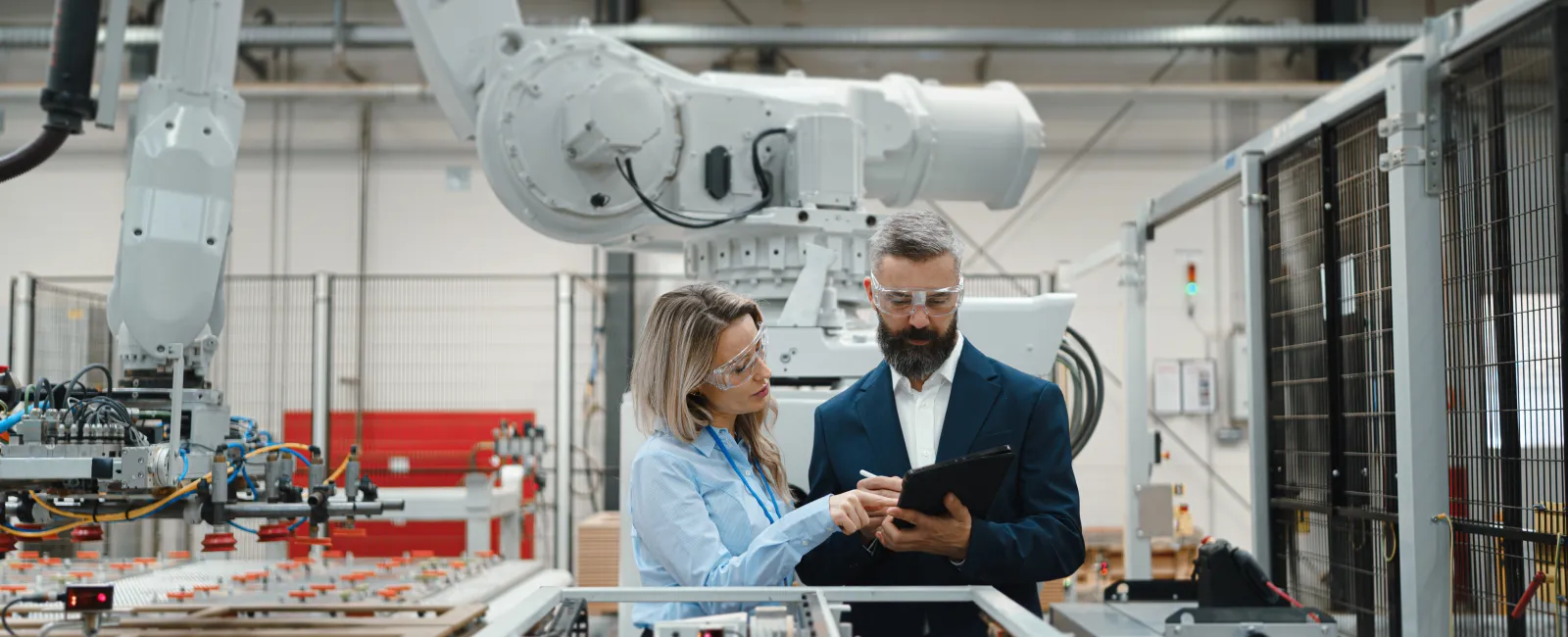 Two engineers in safety glasses discussing a tablet in an advanced factory with robotic machinery and equipment.