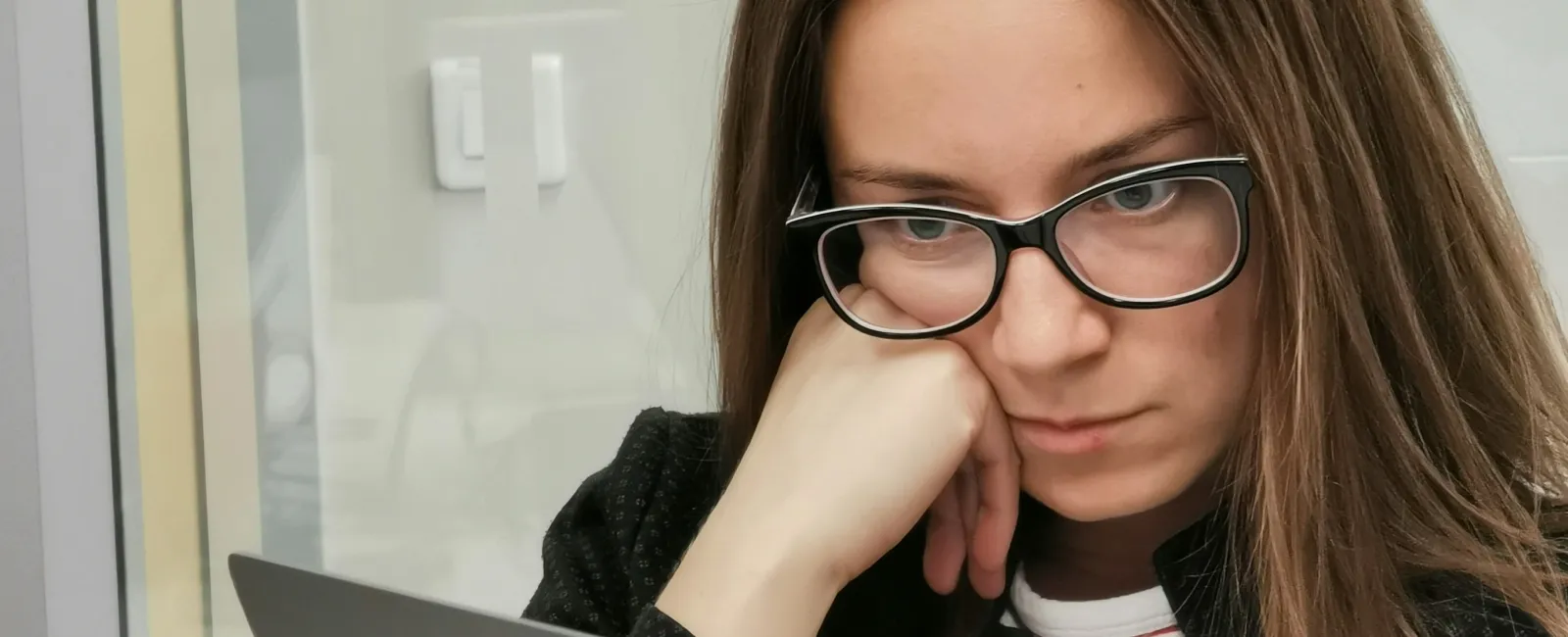Focused woman wearing glasses working on a laptop in an office environment with a notebook and coffee mug nearby.