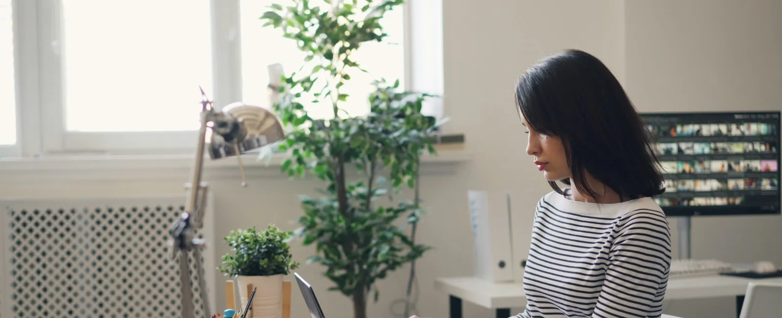 Young woman in striped shirt working on laptop at desk in a bright modern office with plants and books.